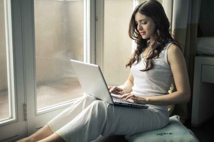 a woman sitting by a window and typing on a laptop